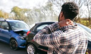 Young man stands by a damaged car, rubbing his neck in pain from a whiplash injury after a traffic accident.
